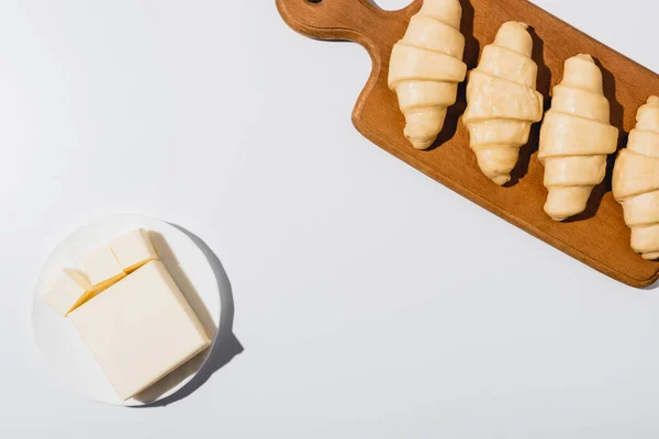 Vista dall'alto di croissant freschi crudi sul tagliere di legno vicino al burro sul piatto con coltello su sfondo bianco — Foto stock