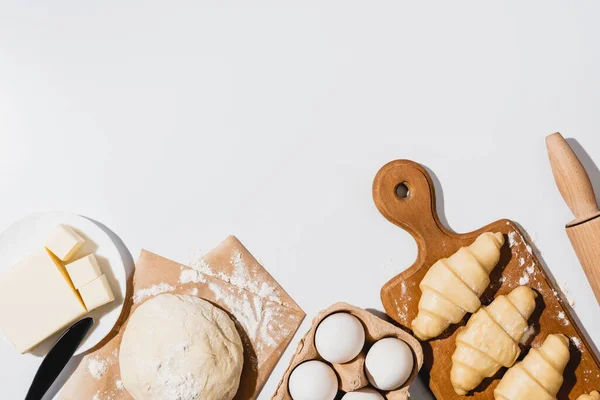 Draufsicht auf frische Croissants auf Holzschneidebrett in der Nähe von rohem Teig, Messer, Nudelholz, Butter und Eiern auf weißem Hintergrund — Stockfoto