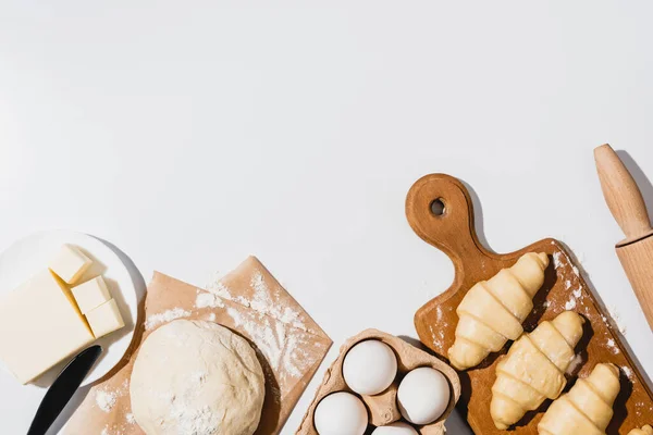 Top view of fresh croissants on wooden cutting board near raw dough, knife, rolling pin, butter and eggs on white background — Stock Photo