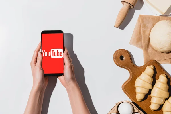 KYIV, UKRAINE - APRIL 22, 2019: partial view of woman holding smartphone with youtube app near raw croissants and ingredients on white background — Stock Photo