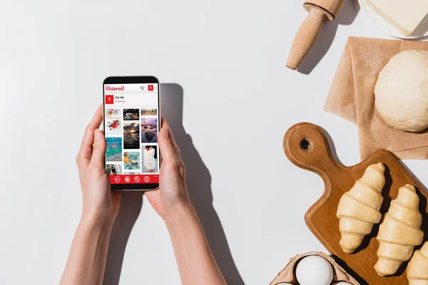 KYIV, UKRAINE - APRIL 22, 2019: partial view of woman holding smartphone with pinterest app near raw croissants and ingredients on white background — Stock Photo