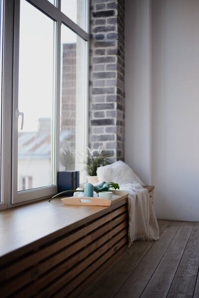  a wide wooden window sill, a large bright window, on a windowsill a wooden tray with a mug, candles. Nearby are books and a green plant in a pot