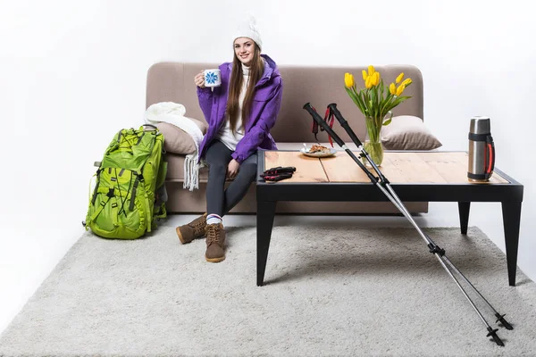 Young woman in warm clothing with backpack and hiking equipment drinking tea at home — Stock Photo