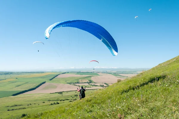 Sporcu bir yamaç paraşütü dağ yamacı üzerinden alır. Arka plan dağlar, alanlar, mavi gökyüzü diğer pilotlar uçan ve