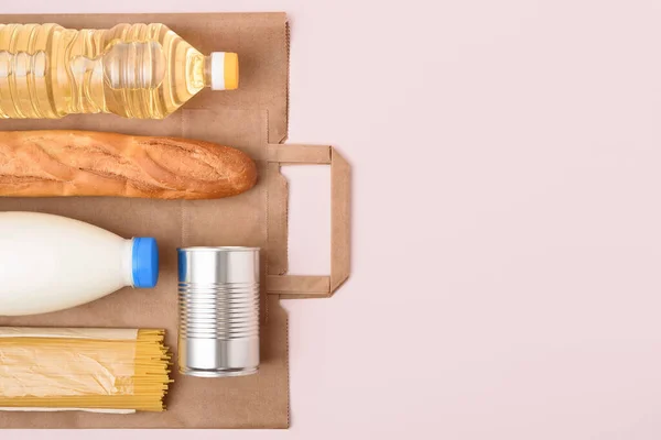Craft package, bottles of sunflower oil and milk, spaghetti, tin can and baguette on the light pink background. Coronavirus safe food delivery. Essential food set. Copy space on the right. Layout.