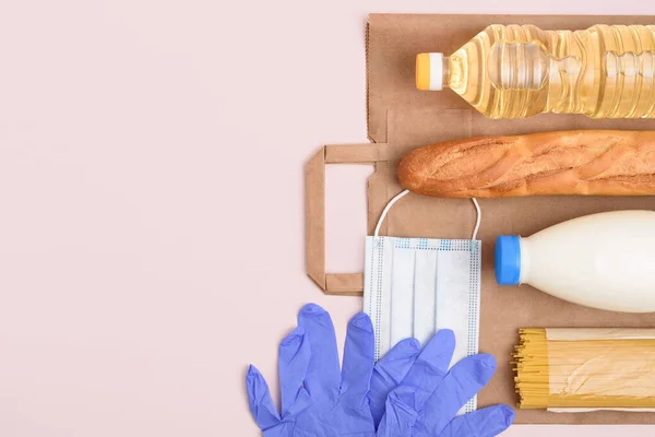 Safe food delivery. Medical mask and gloves, bottles of oil and milk, spaghetti, baguette on the light pink background. Craft package and essential food set. Copy space on the left. Flat lay.