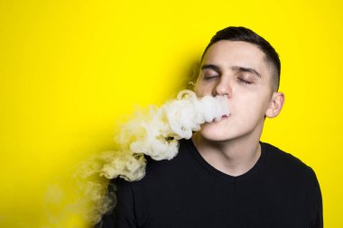 modern saç kesimi ile genç çekici adam elektronik sigara sigara sahiptir ve parlak sarı zıt bir arka plan üzerinde duman bulutları darbeler