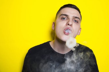 modern bir saç kesimi ile genç çekici adam elektronik sigara sigara sahiptir ve bulutlar ve duman duman halkaları parlak sarı zıt bir arka plan üzerinde darbeler