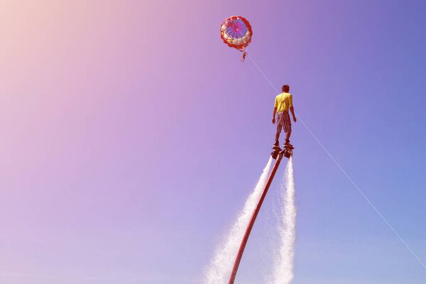 мужчина катается на флайборде на морском пляже с голубым небом и парашютом на заднем плане
