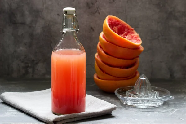 Świeżo wyciskany sok grejpfrutowy w szklanej butelce, sokowirówce i skórkach owoców na konkretnym tle. — Zdjęcie stockowe