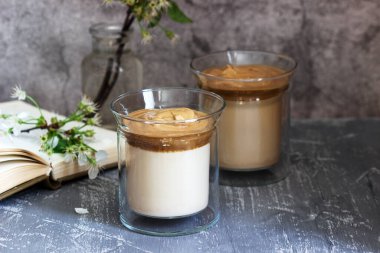 Şeffaf bardaklarda Dalgona Kahve, beton bir masada bir kitap ve çiçekler. Seçici odak.