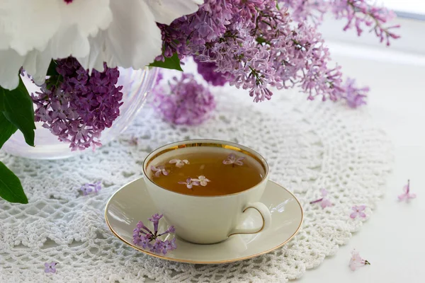 Eine Tasse Kamillentee in einer Porzellantasse und ein Strauß Flieder mit Pfingstrose auf der Fensterbank. — Stockfoto