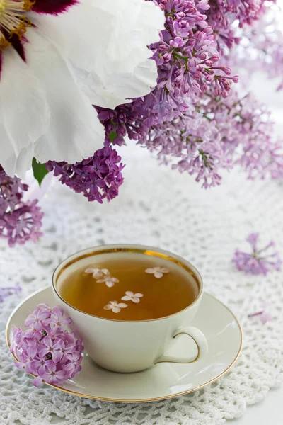 Porselen bir fincanda bir fincan papatya çayı ve pencere kenarında şakayık desenli bir demet leylak..