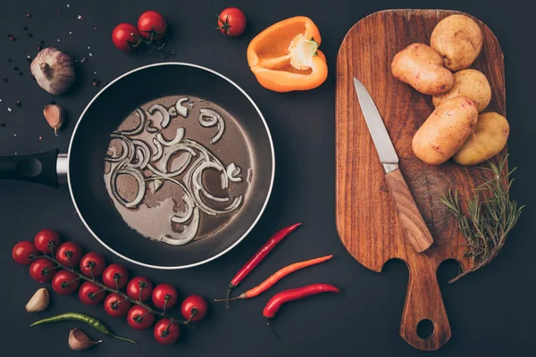 top view of frying pan with onion and wooden board on gray surface