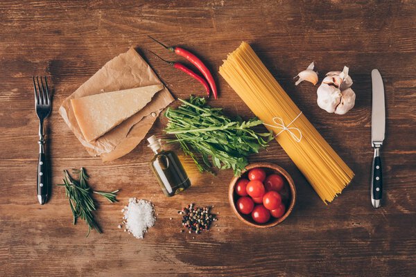 top view of row pasta, fresh ingredients, Parmesan, fork and knife on wooden table