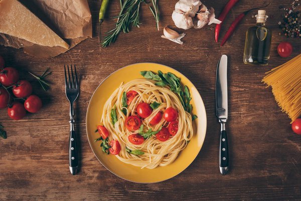 flat lay with traditional italian pasta with tomatoes and arugula in plate on wooden table with ingredients