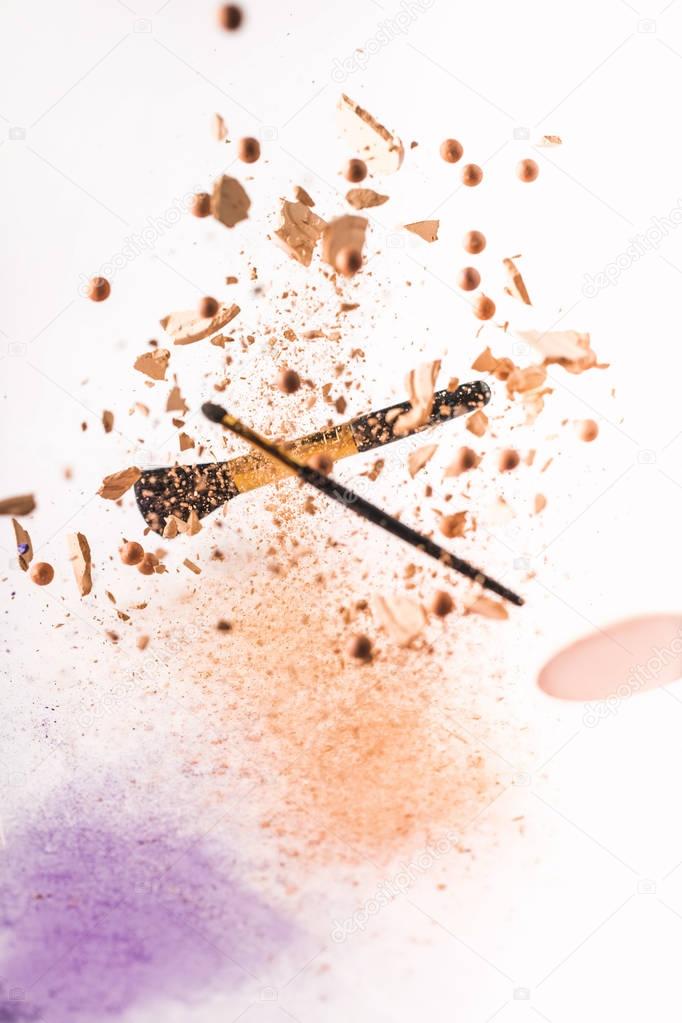 crushed cosmetic powder with makeup brushes falling isolated on white