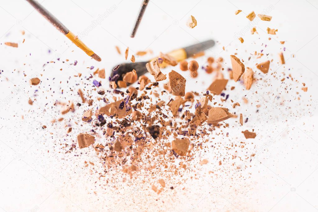 close-up shot of pieces of cosmetic powder with makeup brushes falling isolated on white