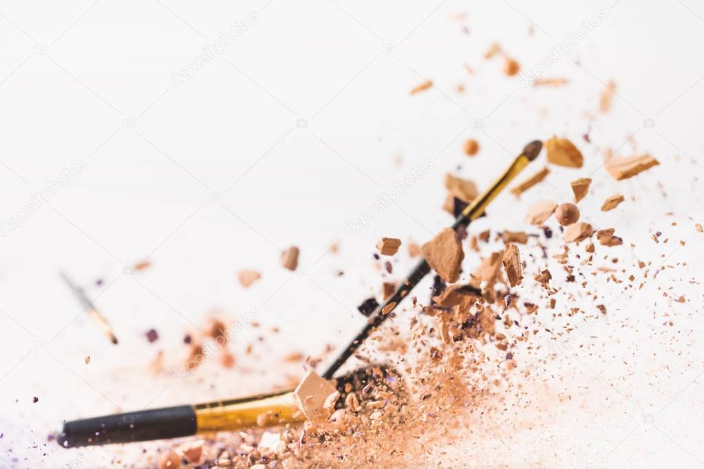 pieces of cosmetic powder with makeup brushes falling isolated on white