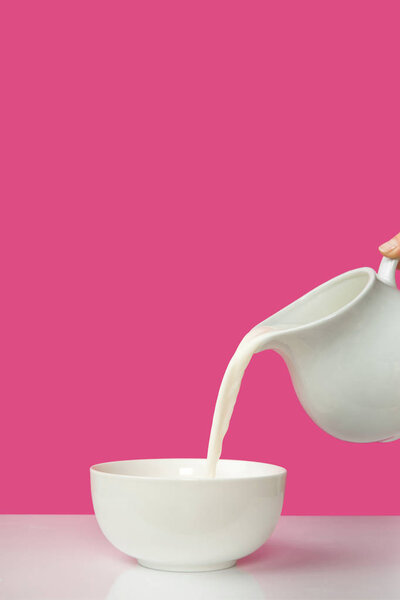 cropped shot of hand pouring fresh milk from jug to bowl on pink      