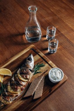 close-up shot of grilled fish with lemon on wooden board with vodka in decanter and shots clipart