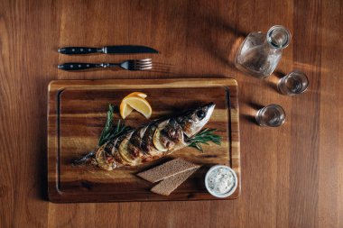 top view of grilled fish with lemon on wooden board with vodka in decanter and shots clipart