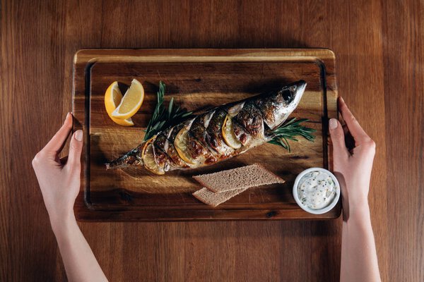 cropped shot of woman holding grilled fish with lemon on wooden board