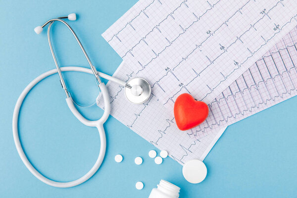 стетоскоп, бумага с кардиограммой, разбросанные таблетки и красное сердце изолированы на синем фоне
  