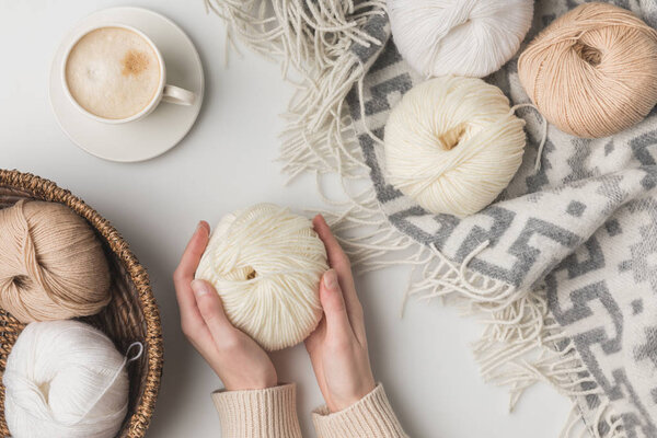 cropped view of woman holding cup of coffee and yarn balls on blaket and in basket on white background 