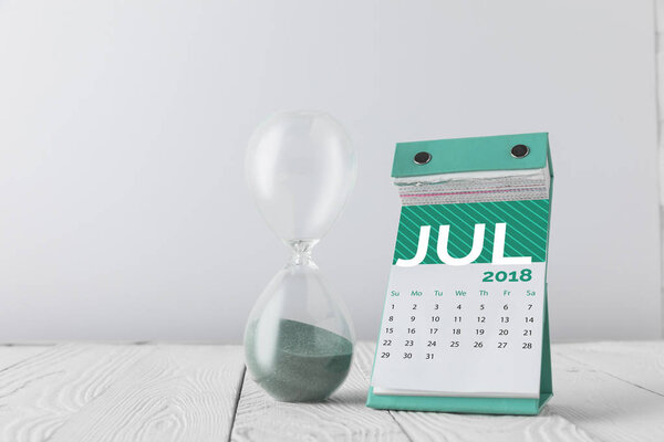 close up view of hourglass and july calendar on wooden tabletop isolated on white