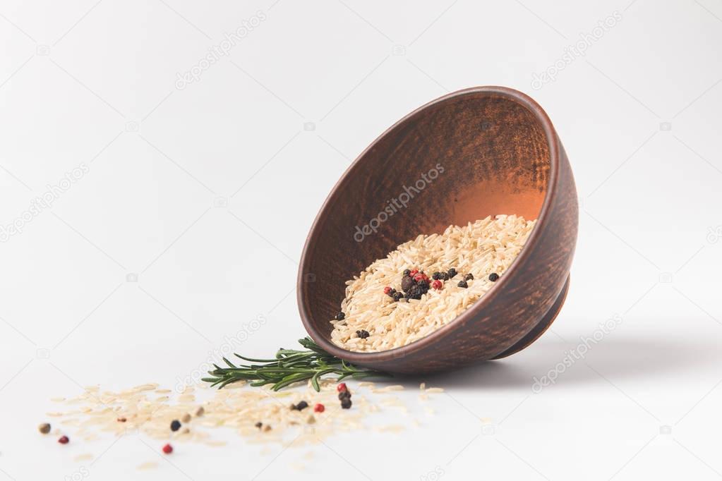 raw rice and peppercorns spilling out bowl on white surface