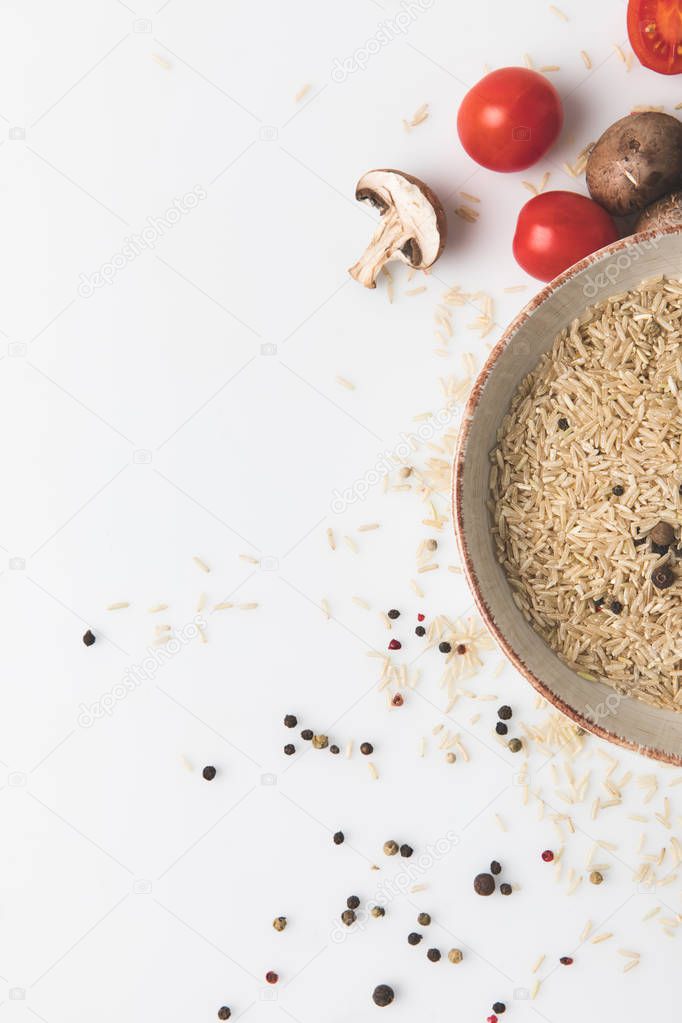 top view of raw rice and spices in bowl with mushrooms and tomatoes on white surface with copy space