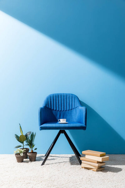 голубое кресло с чашкой кофе и книгами с цветочными горшками на полу перед синей стеной
