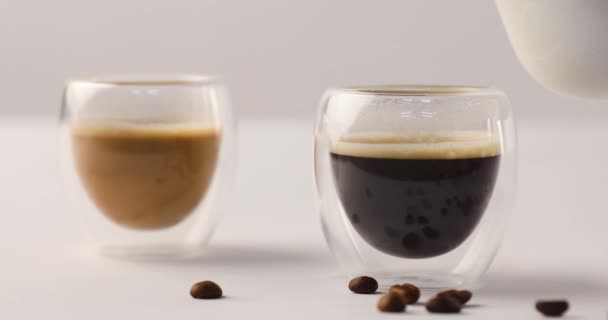 Pouring milk from jug in one of two cups with coffee on white background with coffee beans
