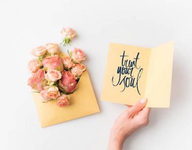 hand holding paper with TRUST YOUR SOUL sign beside pink flowers in envelope isolated on white clipart