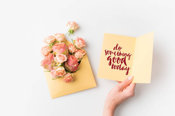 hand holding paper with DO SOMETHING GOOD TODAY inscription beside pink flowers in envelope isolated on white