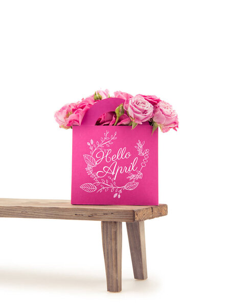 Крупный план нежных цветущих розовых цветов в розовом бумажном пакете с надписью HELLO APRIL на деревянной скамейке, изолированной на белом
