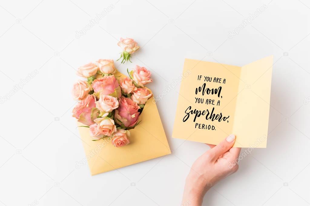 hand holding paper with mothers day phrase beside pink flowers in envelope isolated on white
