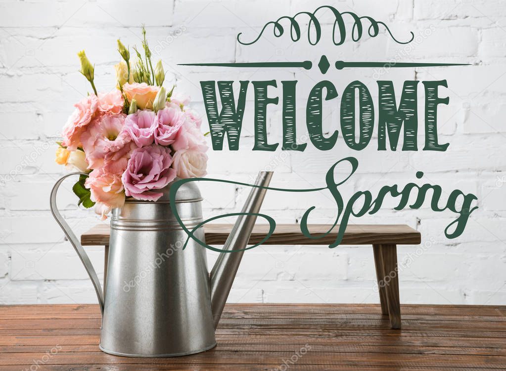 Beautiful blooming flowers in watering can and small bench on wooden surface with WELCOME SPRING lettering