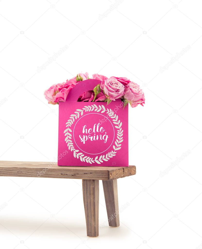 Close-up view of tender blooming rose flowers in pink paper bag with HELLO SPRING lettering on wooden bench isolated on white