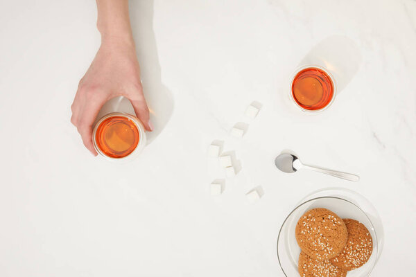 cropped shot of human hand with cup of tea and tasty oatmeal cookies on grey