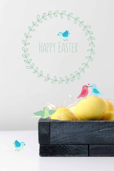 yellow painted easter eggs in wooden box with drawn birds and happy easter lettering on white table