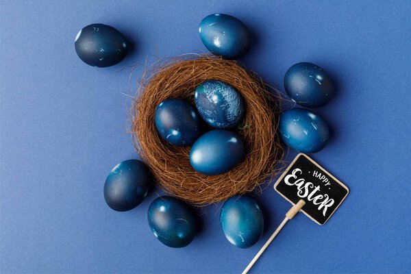 top view of blue painted easter eggs in decorative nest and happy easter lettering on signboard on blue surface