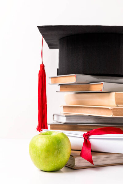 Apple near diploma and pile of books with academic cap on top isolated on white