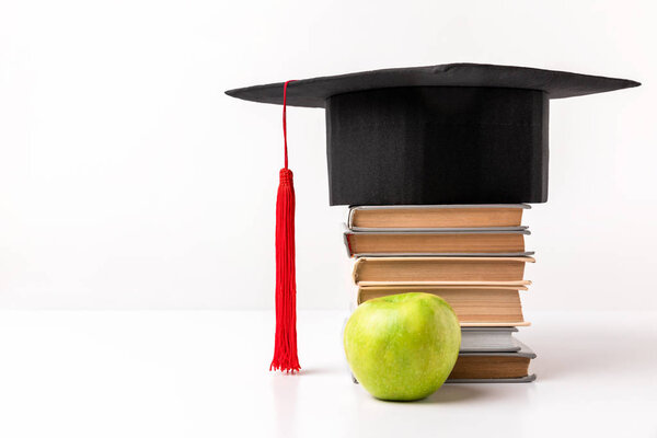 Close up view of apple near pile of books with academic cap on top isolated on white