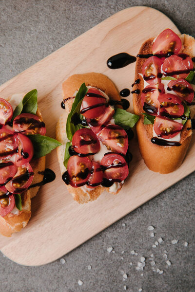 верхний вид вкусной брускетты с помидорами и бальзамическим на деревянной доске на сером
 