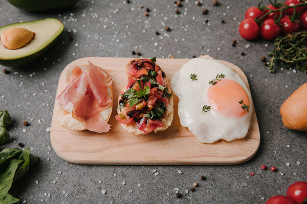 antipasto bruschetta and fried egg on wooden cutting board and ingredients on grey  