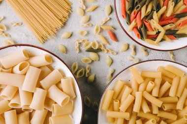 top view of different raw pasta in bowls on messy concrete surface  clipart