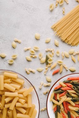 top view of different raw pasta in bowls on messy concrete tabletop clipart