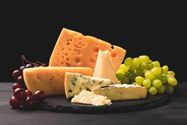 Closeup shot of different types of cheese on board with grapes on black 